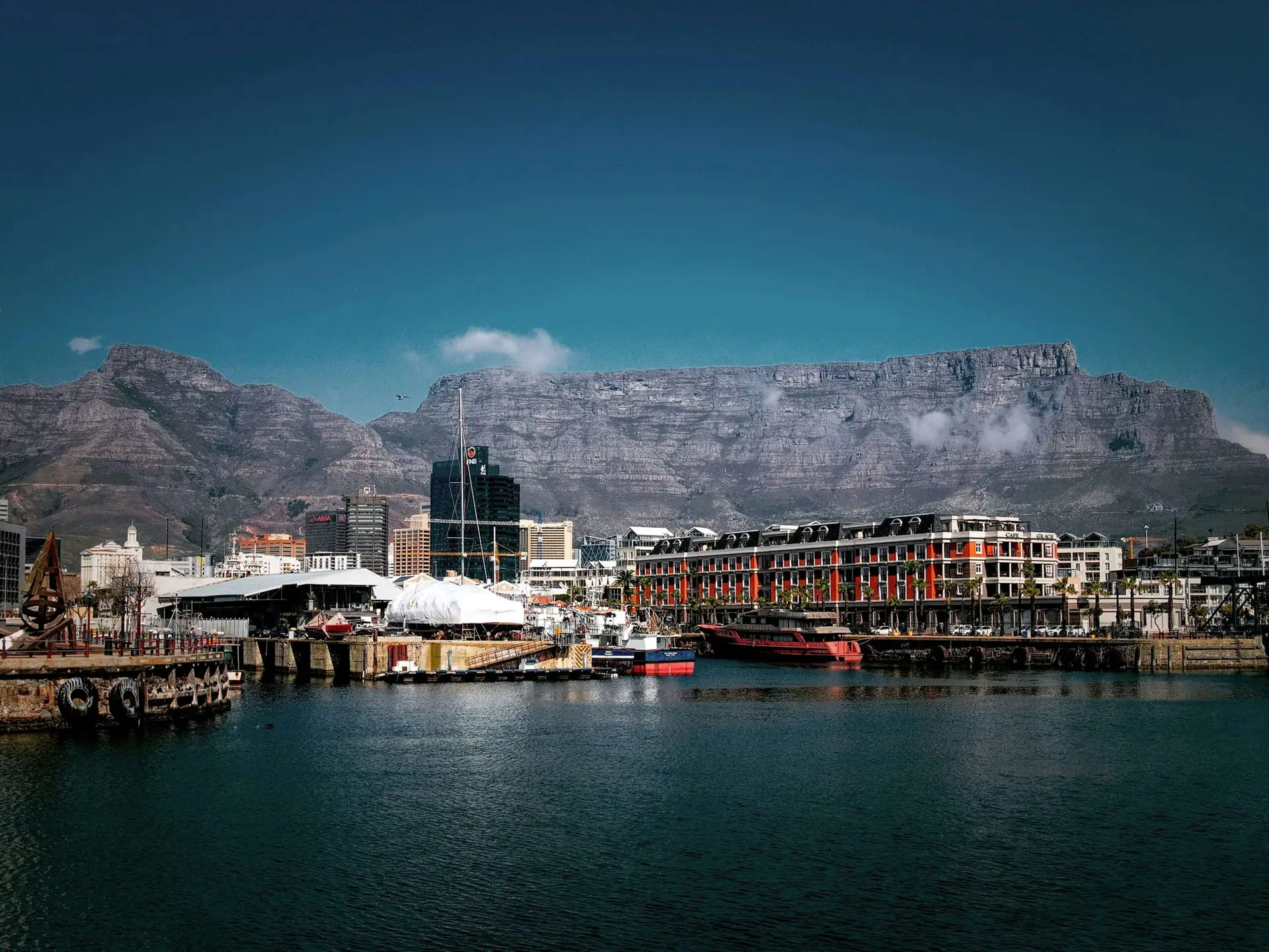 Panoramic view of the Victoria & Alfred Waterfront in Cape Town, South Africa, with Table Mountain in the background. Red and blue boats are docked in the calm harbor, modern buildings line the promenade, and the iconic flat-topped mountain rises under a clear blue sky formatted for our provisions