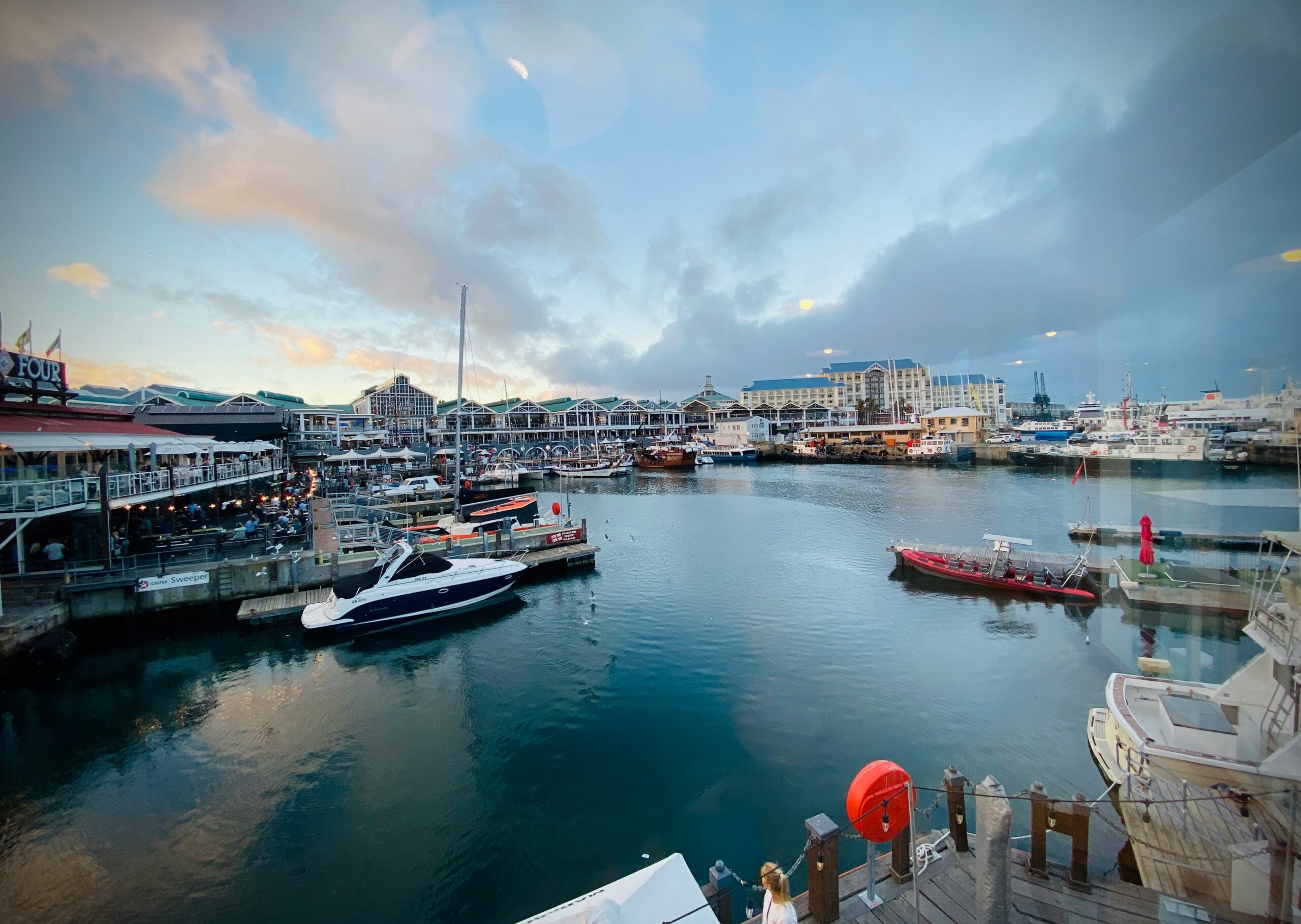 Twilight view of the bustling Victoria & Alfred Waterfront in Cape Town, South Africa. Numerous boats and yachts are docked in the harbor, with people enjoying outdoor dining and promenades along the waterfront, under a colorful evening sky.